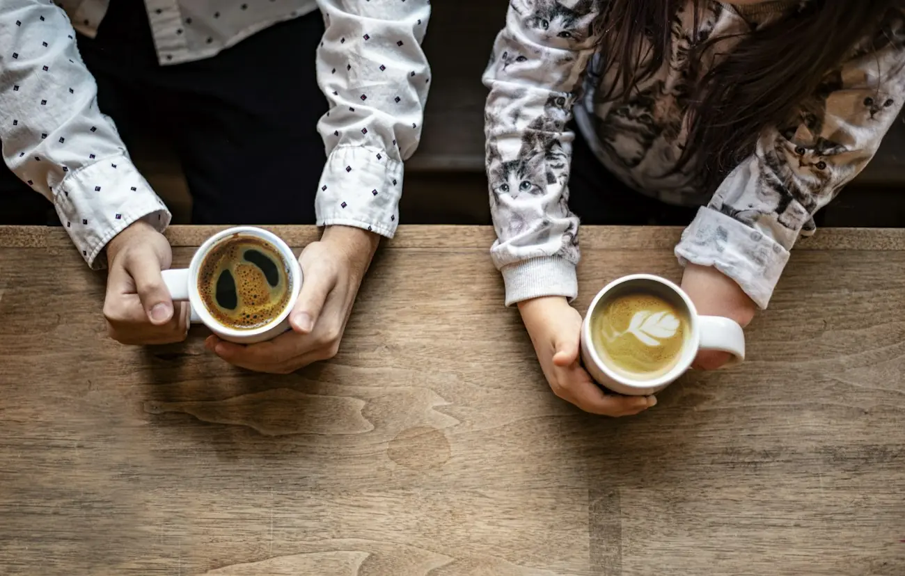 Two people holding cups of coffee across a wooden cafe table